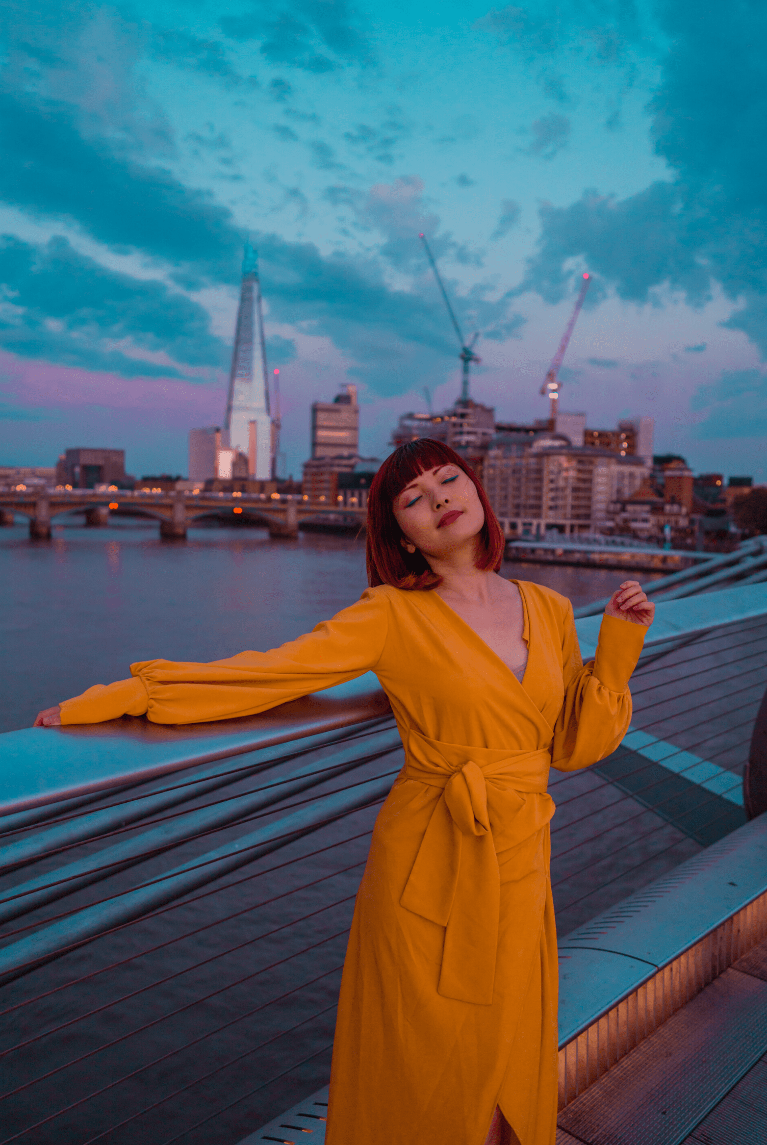 A woman wearing the Sarvin Mustard Wrap Maxi Dress with balloon bishop sleeves stands by a bridge railing at dusk, city skyline and river behind her. The partly cloudy sky glows in blue and purple hues, cranes visible in the distance.