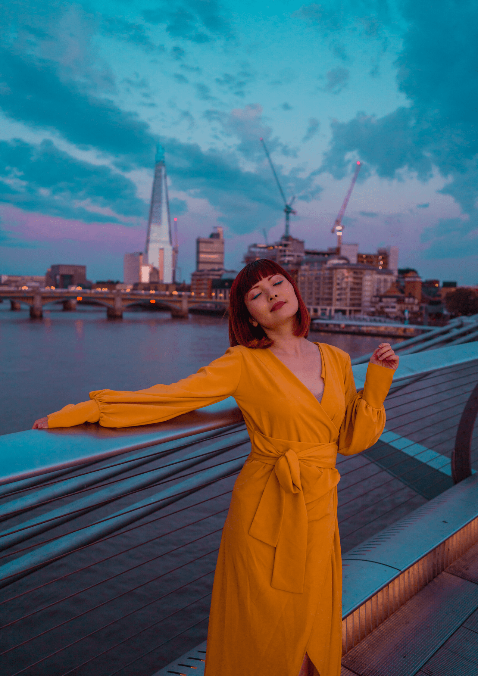 A woman wearing the Sarvin Mustard Wrap Maxi Dress with balloon bishop sleeves stands by a bridge railing at dusk, city skyline and river behind her. The partly cloudy sky glows in blue and purple hues, cranes visible in the distance.