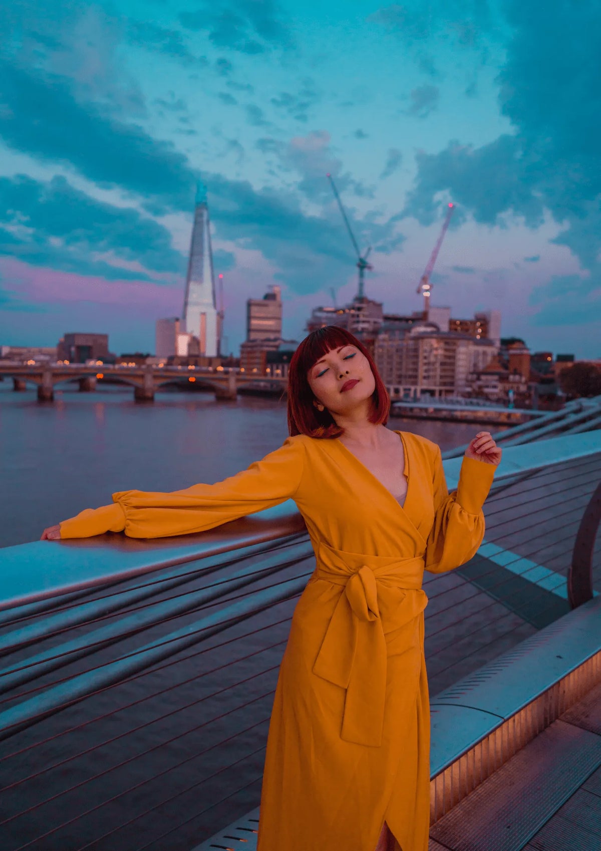 A woman wearing the Sarvin Mustard Wrap Maxi Dress with balloon bishop sleeves stands by a bridge railing at dusk, city skyline and river behind her. The partly cloudy sky glows in blue and purple hues, cranes visible in the distance.