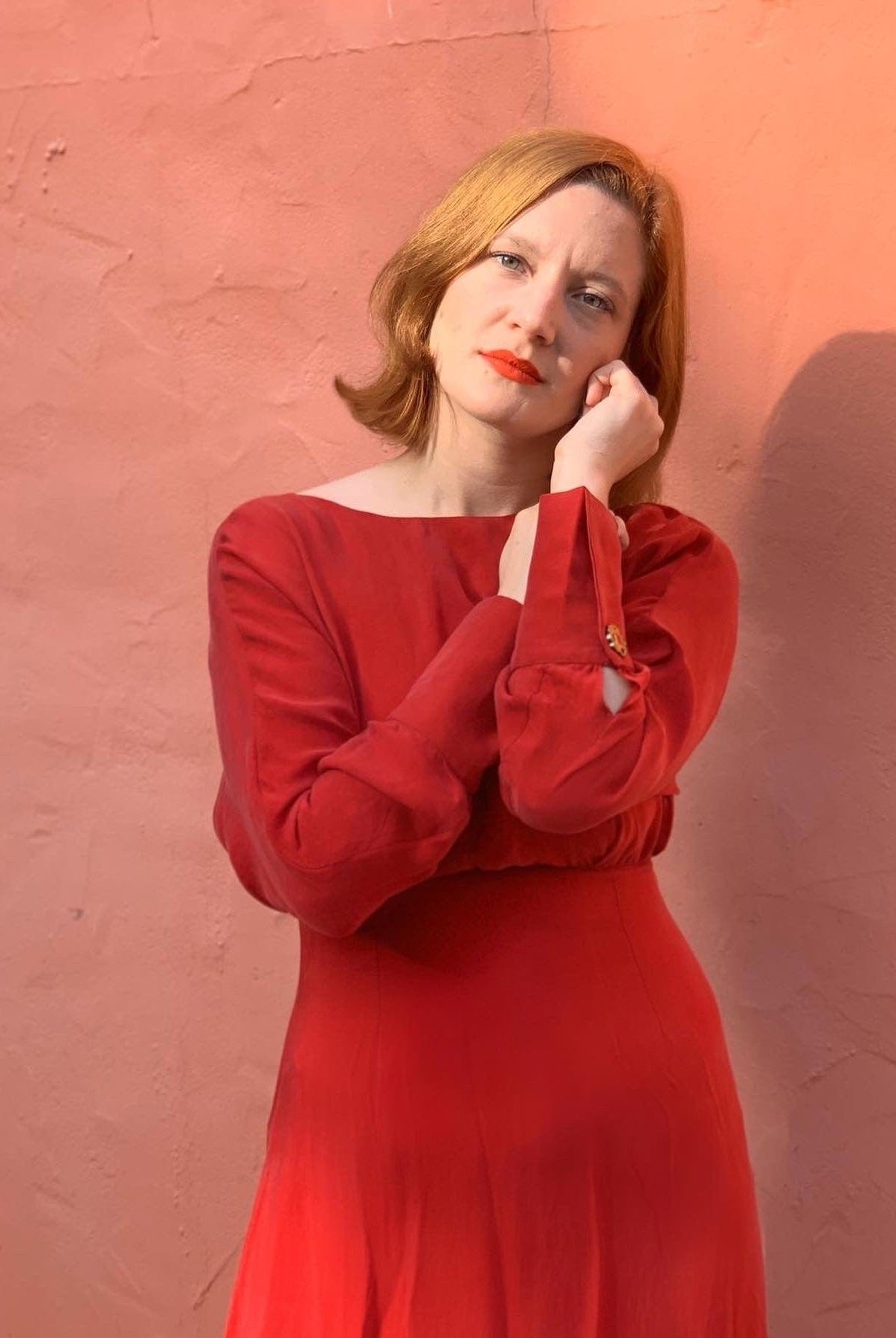 A woman with light skin and short, light brown hair poses against a peach wall, wearing the Sarvin Long Sleeve Red Midi Dress and resting her face on her hand while facing the camera.