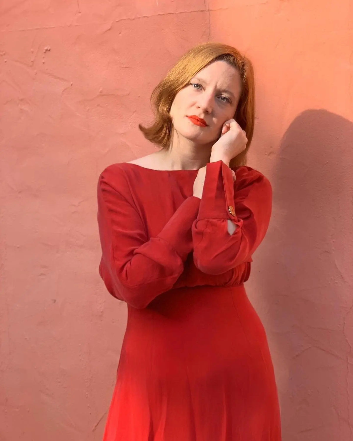 A woman with light skin and short, light brown hair poses against a peach wall, wearing the Sarvin Long Sleeve Red Midi Dress and resting her face on her hand while facing the camera.