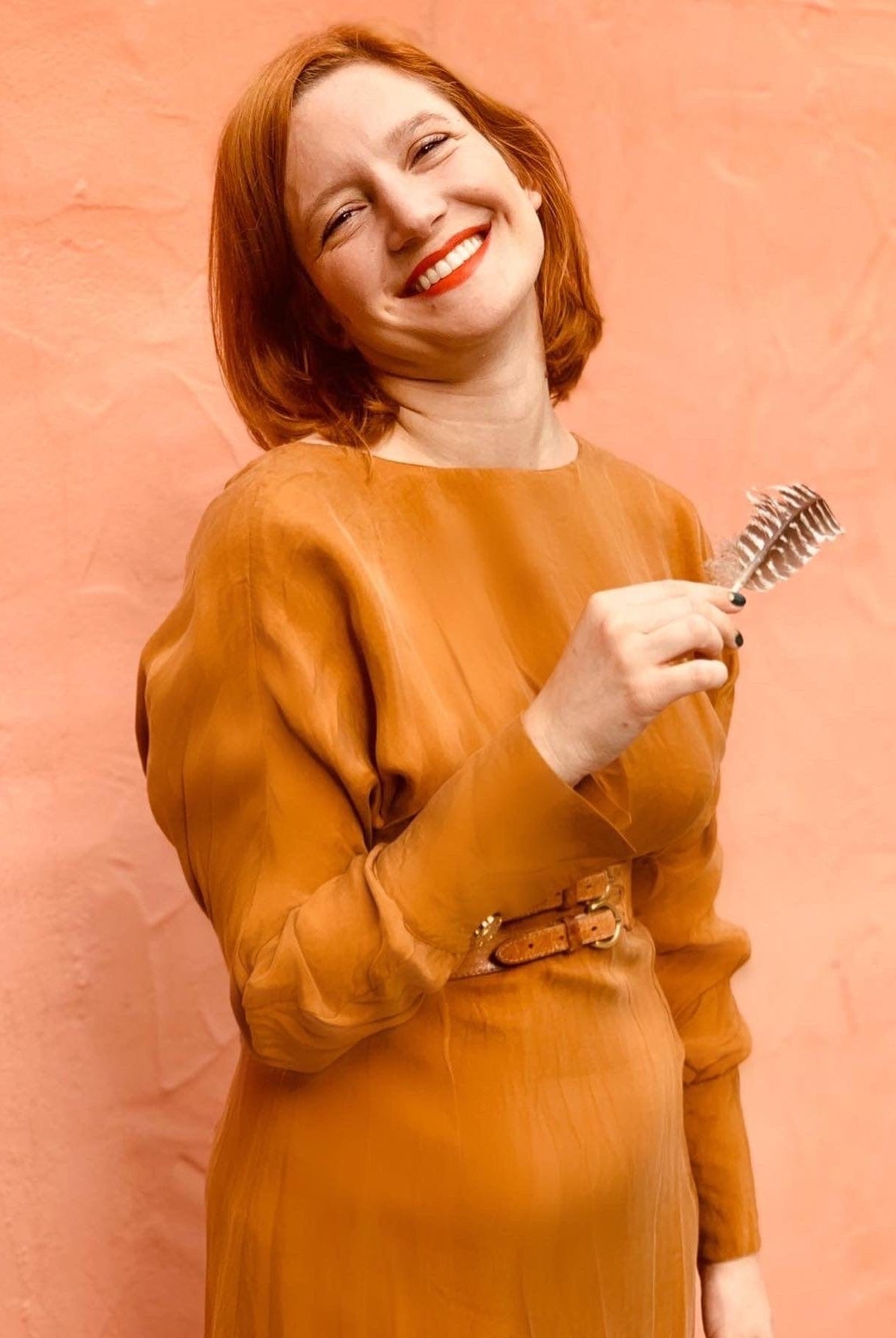 A smiling person with short red hair wears the Sarvin Mustard Yellow Midi Dress, styled with a belt, and holds a metal hair clip. The background is a plain peach-colored wall.