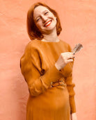 A smiling person with short red hair wears the Sarvin Mustard Yellow Midi Dress, styled with a belt, and holds a metal hair clip. The background is a plain peach-colored wall.