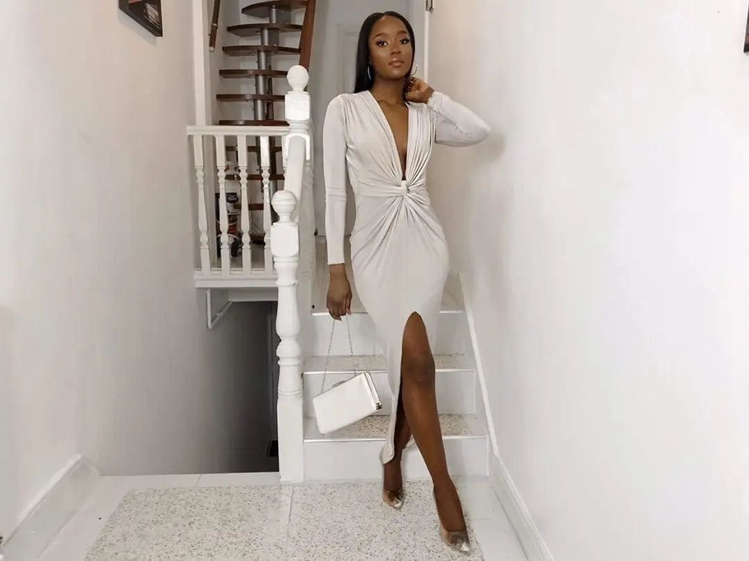 A woman wearing the Sarvin Low Cut Midi Dress stands on indoor stairs, holding a white handbag. She completes her look with transparent pointed heels, and a spiral staircase is visible behind her.