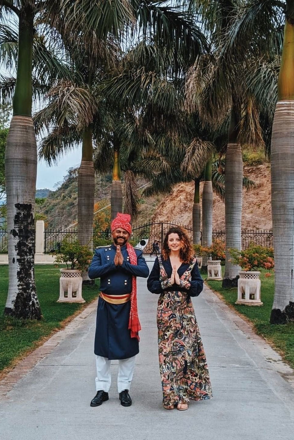 A man in traditional Indian attire and a woman in a Sarvin Hand painted Long Sleeve Maxi Dress stand side by side on a walkway, palms pressed together in namaste amid palm trees and lush greenery.