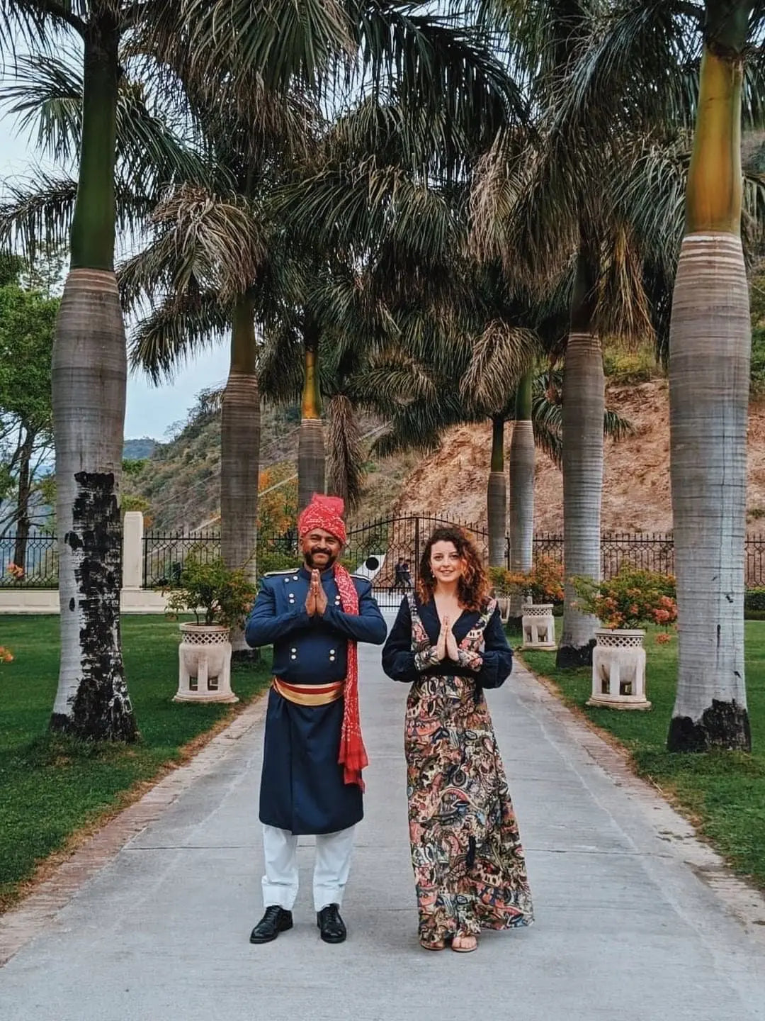 A man in traditional Indian attire and a woman in a Sarvin Hand painted Long Sleeve Maxi Dress stand side by side on a walkway, palms pressed together in namaste amid palm trees and lush greenery.