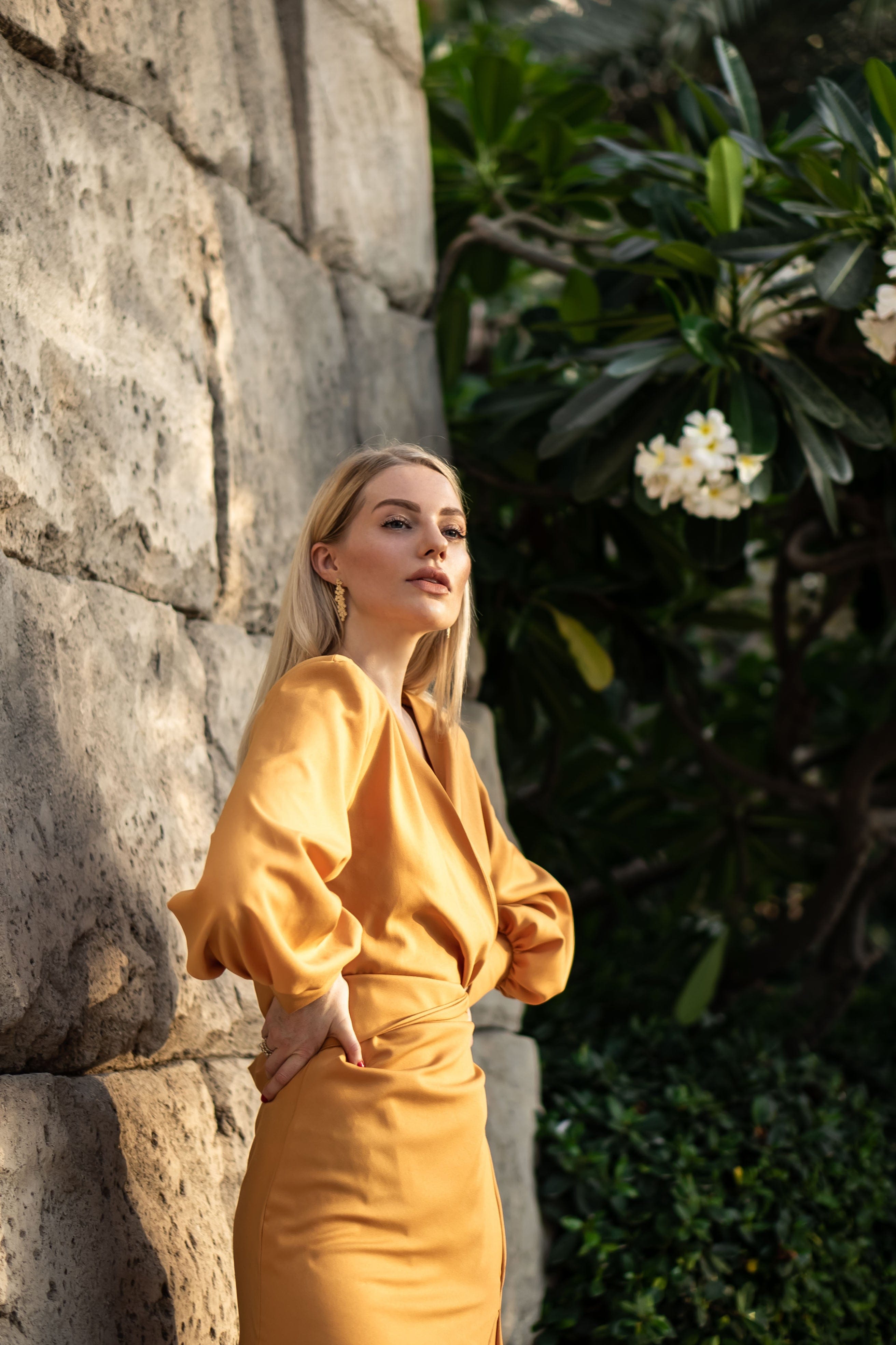 A woman with blonde hair stands near a stone wall and white flowers, wearing the Sarvin Mustard Wrap Maxi Dress with vintage-inspired satin and balloon bishop sleeves. She gazes upward with one hand on her hip.