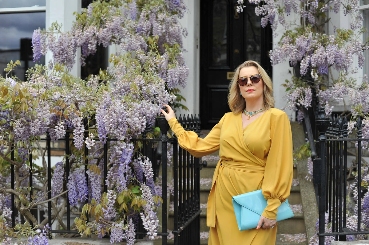 A woman in the Sarvin Mustard Wrap Maxi Dress with balloon bishop sleeves and sunglasses stands by a black iron fence draped with purple wisteria, holding a blue clutch and looking ahead.