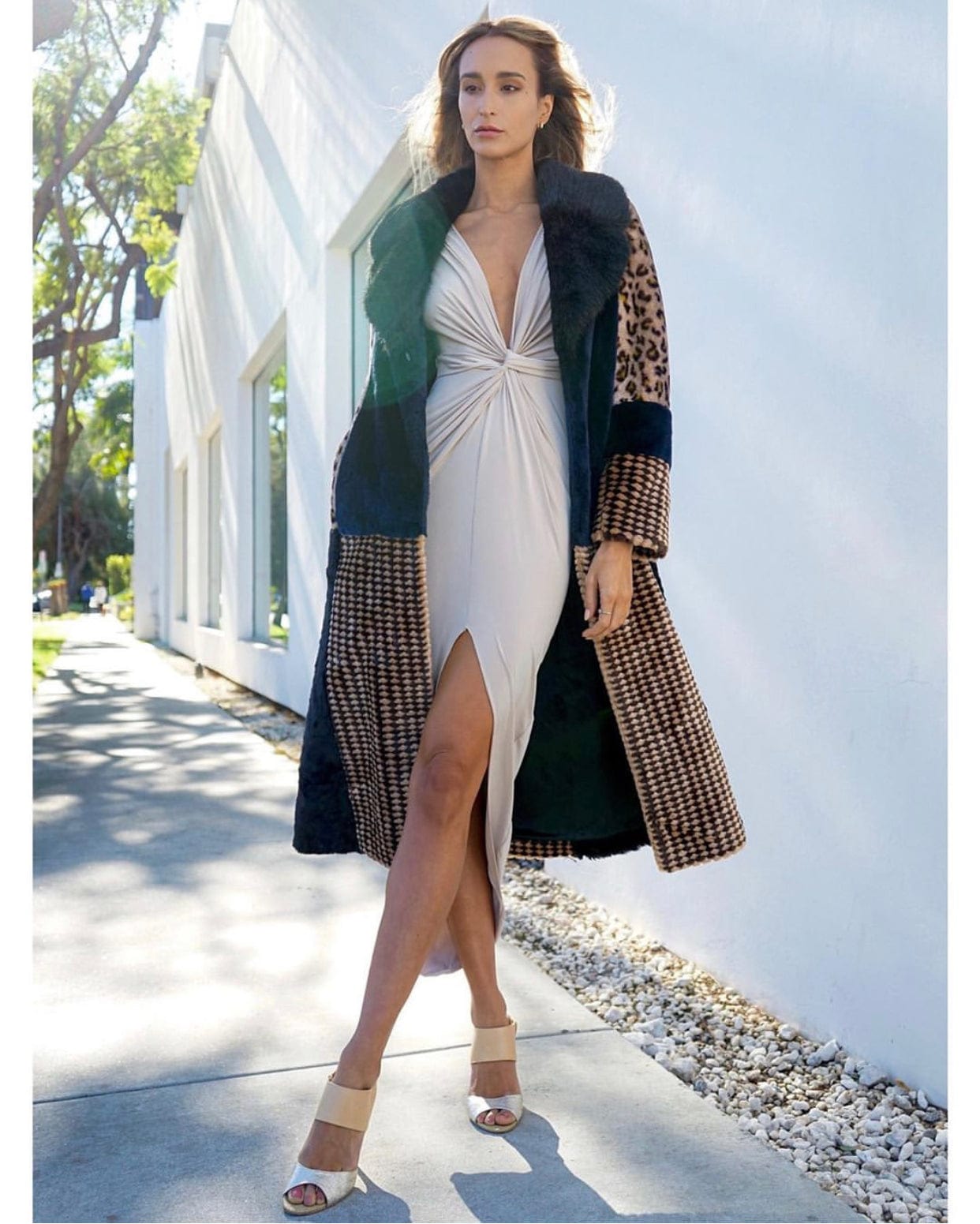 A woman strolls by a modern white building on a sunny day, wearing the Sarvin Low Cut Midi Dress with a high slit under a long patterned coat with fur trim, paired with beige heeled sandals.