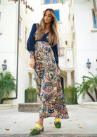 A woman with long hair stands outside a building wearing the Sarvin Hand painted Long Sleeve Maxi Dress and green shoes, with palm trees and architectural details in the background.