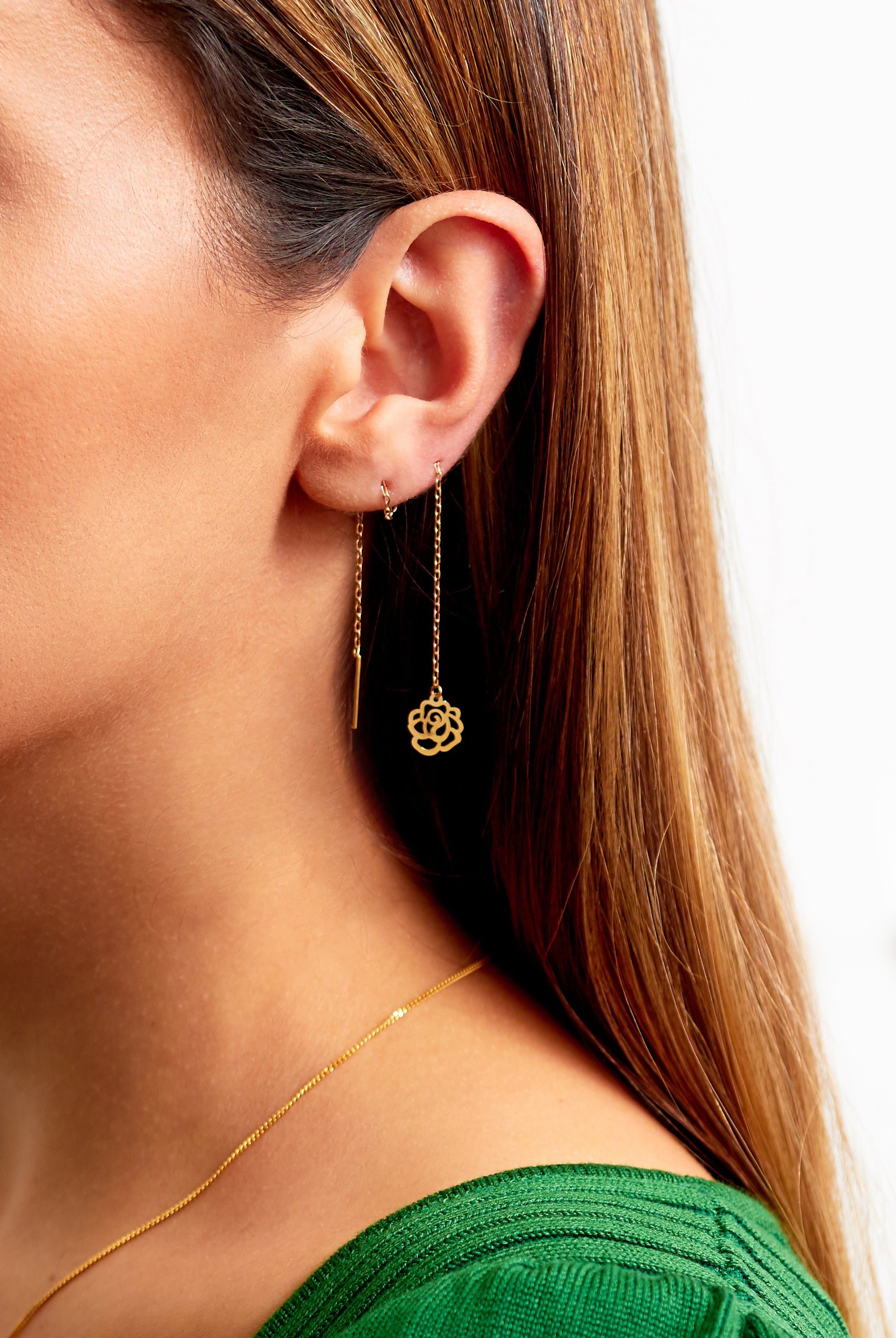 Close-up of a woman wearing Sarvin Flower Shape 18K Gold Drop Earrings—one is a bar, the other a pull-through with a flower pendant. She has straight brown hair, a green top, and a thin gold necklace.