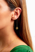 Close-up of a woman wearing Sarvin Flower Shape 18K Gold Drop Earrings—one is a bar, the other a pull-through with a flower pendant. She has straight brown hair, a green top, and a thin gold necklace.