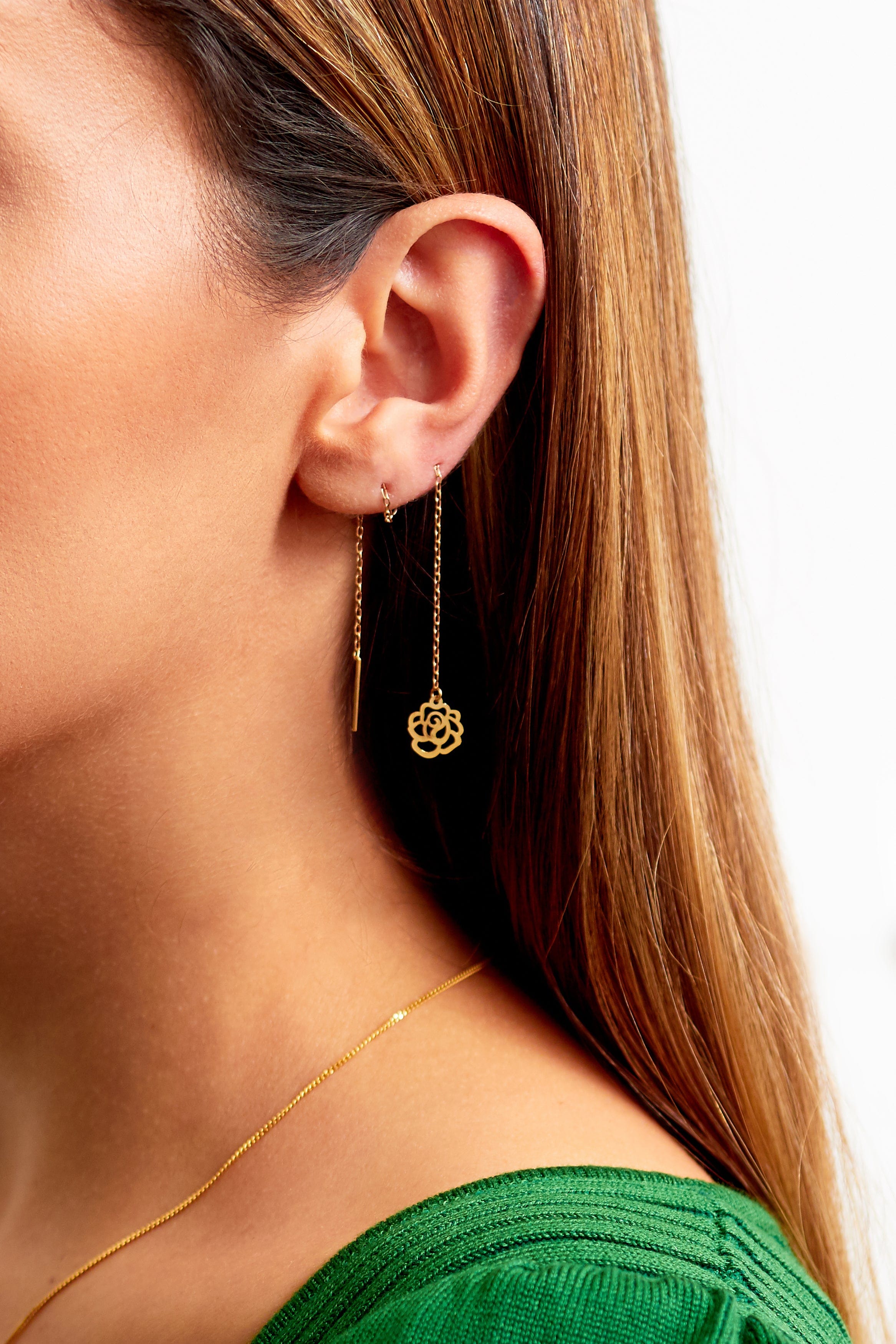 Close-up of a woman wearing Sarvin Flower Shape 18K Gold Drop Earrings—one is a bar, the other a pull-through with a flower pendant. She has straight brown hair, a green top, and a thin gold necklace.