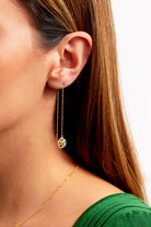 Close-up of a woman's ear wearing Sarvin's Flower Shape 18K Gold Drop Earrings with a delicate chain and circular design. She has straight brown hair, a green top, and a thin gold necklace, set against a plain white background.