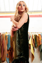 A woman with long blonde hair wears the Sarvin Silk Velvet Feather Trim Dress in forest green, standing indoors before colorful hanging fabrics and gazing slightly up and left with one arm bent across her body.