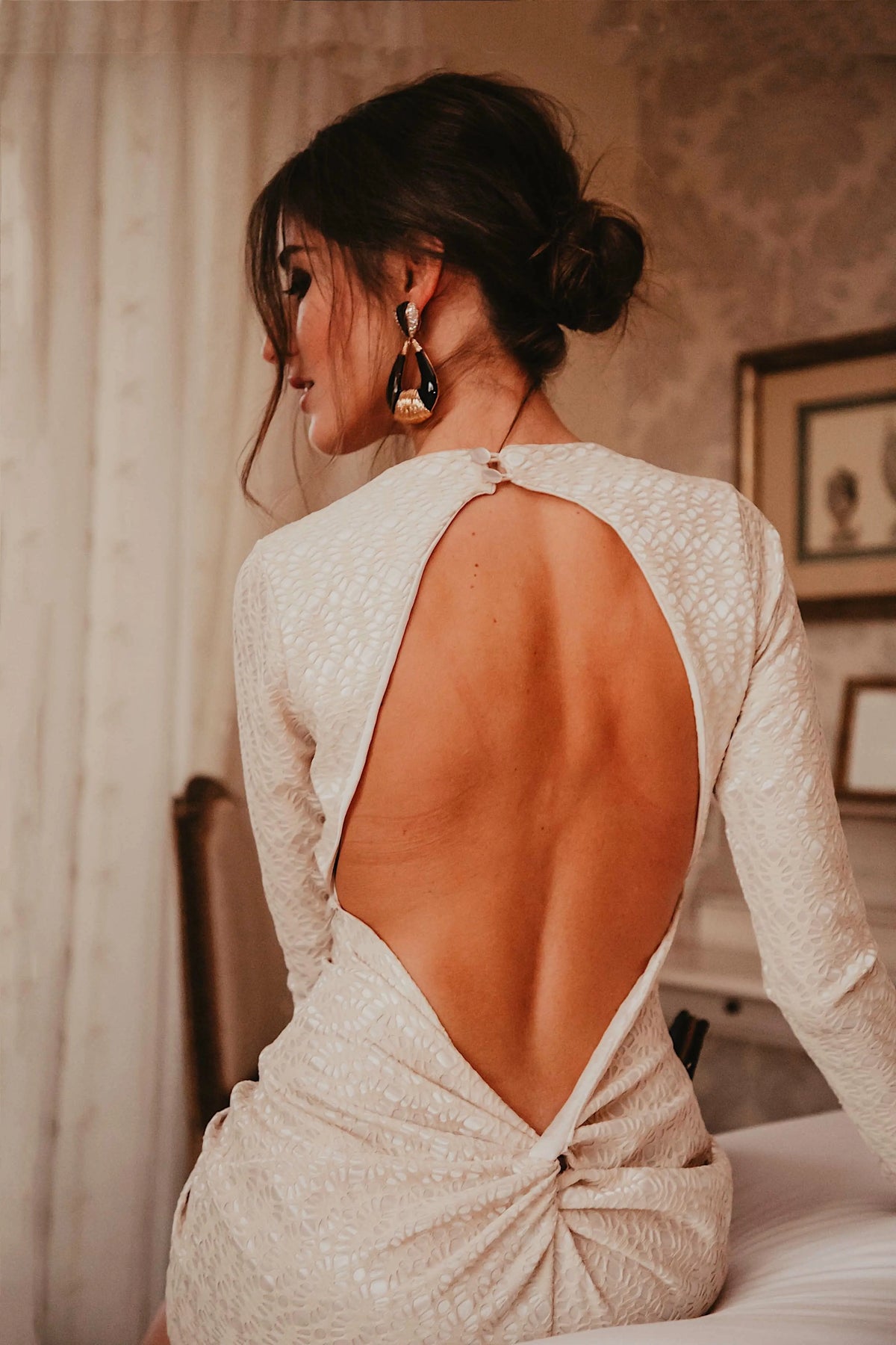 A woman with dark hair in an updo sits on a bed in a softly lit room, wearing the Sarvin Ivory Lace Backless Dress, its delicate open-back detail elegantly highlighted as she faces away from the camera.
