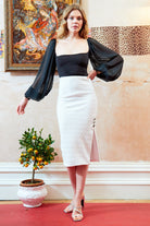 A woman wears the Sarvin Black Mesh Bodysuit with sheer billowy sleeves and a white pencil skirt, standing barefoot on a red carpet beside a potted orange tree, against leopard print wallpaper and a painting.