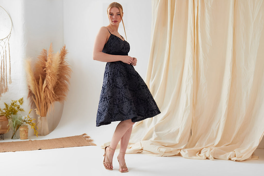 blonde woman with hair in pony-tail wearing a navy jaquard 60s inspired skater dress and nude heels, in a studio