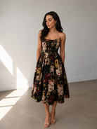 A woman stands indoors wearing the Sarvin Golshifteh Floral Corset Midi Dress with her hands in the pockets, long dark hair down, and looking slightly downward.