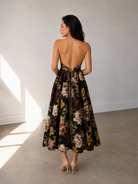 A woman stands indoors on a bare floor, facing away from the camera, wearing the Sarvin Golshifteh Floral Corset Midi Dress and nude high heels. The background and lighting are neutral and minimal.