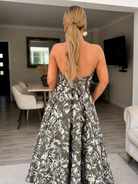 A woman with long blonde hair in a low ponytail stands indoors, facing away, wearing the Sarvin Tala Corset Midi Dress—a strapless black, white, and gray floral piece—in a modern living and dining area.