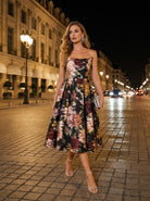 A woman in the Sarvin Golshifteh Floral Corset Midi Dress and heels walks on a cobblestone street at night, holding a small clutch, her pleated skirt swaying as she passes historic buildings lit by streetlights.
