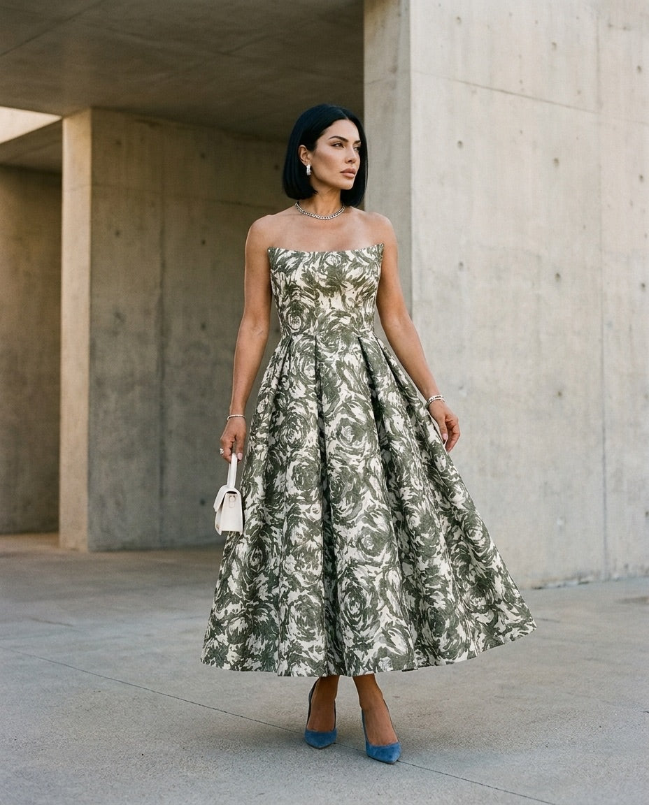 A woman stands outdoors against a concrete wall, wearing the Sarvin Tala Corset Midi Dress, styled with straight black hair, jewelry, blue high heels, and holding a small white purse.