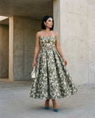 A woman stands outdoors against a concrete wall, wearing the Sarvin Tala Corset Midi Dress, styled with straight black hair, jewelry, blue high heels, and holding a small white purse.