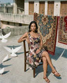 A woman in the Sarvin Golha Floral Corset Dress sits on a wooden chair on a rooftop, with patterned rugs hanging behind her and white doves gathered nearby and flying overhead.