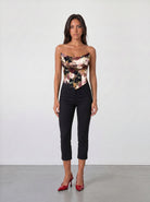 A woman with long dark hair faces forward against a plain light wall, wearing the Sarvin Ivy Corset Top with a waist-cinching floral print, paired with black cropped pants and red high heels. Her expression is neutral.