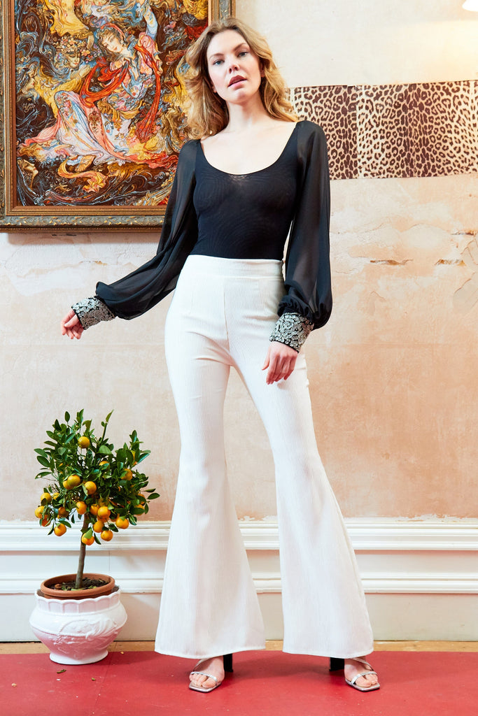 A woman is standing in a room wearing white pants and a Sarvin Black Mesh Bodysuit Long Sleeve.